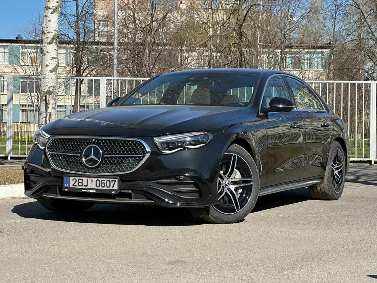 سيارة Mercedes E-Class سيدان سوداء مصورة في موقف خارجي أمام سياج وأشجار ومبنى